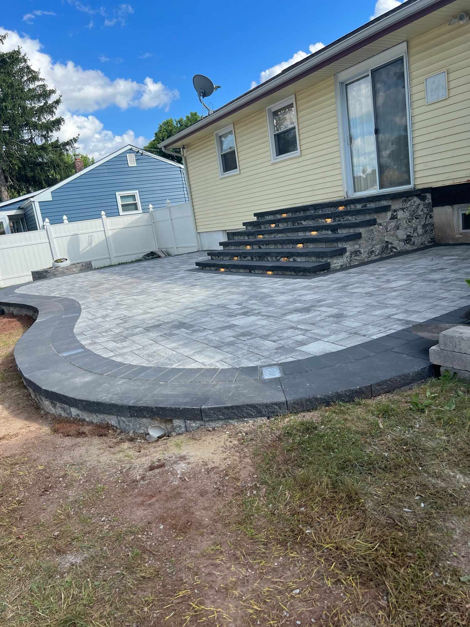 Curved paver patio with charcoal border and stone-veneer steps at the back of a yellow New Jersey home