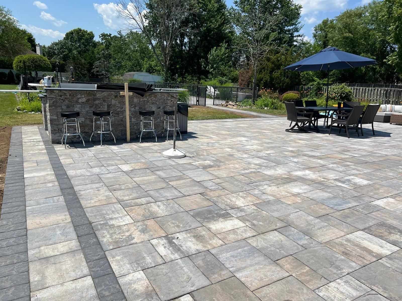 Large paver patio with a stone outdoor kitchen bar, bar stools and dining set, built by Mana Concrete in Central New Jersey