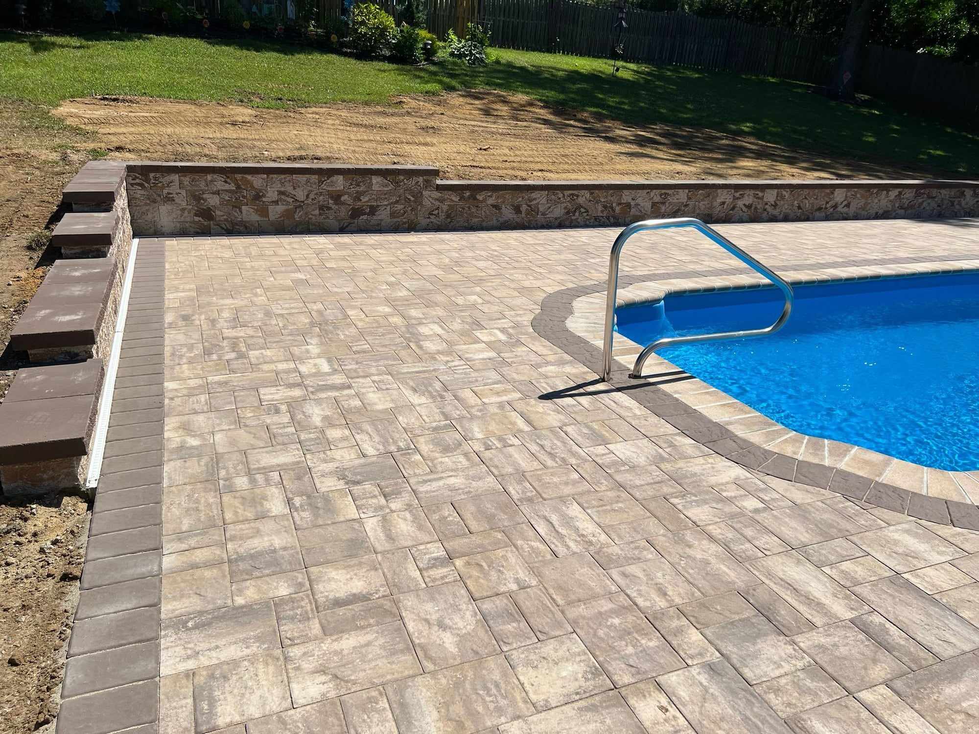 Tan paver pool deck with a block retaining wall along the slope of a New Jersey backyard